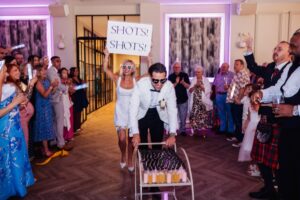 A couple in wedding attire pushes a trolley filled with shot glasses as guests cheer and clap in a warm, festive indoor setting at Woodlands Hotel, Leeds