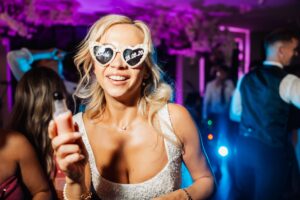 A bride in a white beaded dress smiles and holds a drink at a lively wedding reception with purple and blue stage lighting