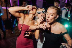 Three women in elegant dresses dance and drink champagne at a lively party, with vibrant stage lighting and other guests in the background at Woodlands Hotel, Leeds