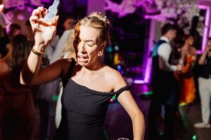 A woman in a black dress dances energetically at a wedding reception, holding a small bottle in her raised hand