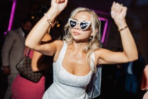 A woman in a silver sequined dress dances with her arms raised, wearing heart-shaped sunglasses with 'Bride' and a date written on them at Woodlands Hotel, Leeds