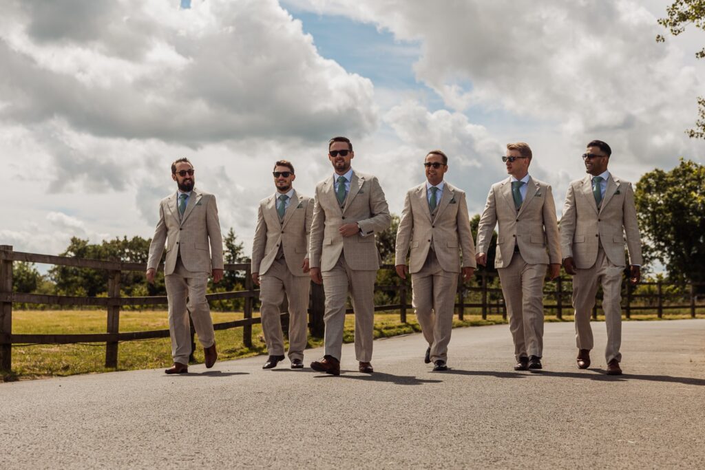 Six men in beige suits and sunglasses walk along a paved path, with a wooden fence and greenery in the background under a partly cloudy sky