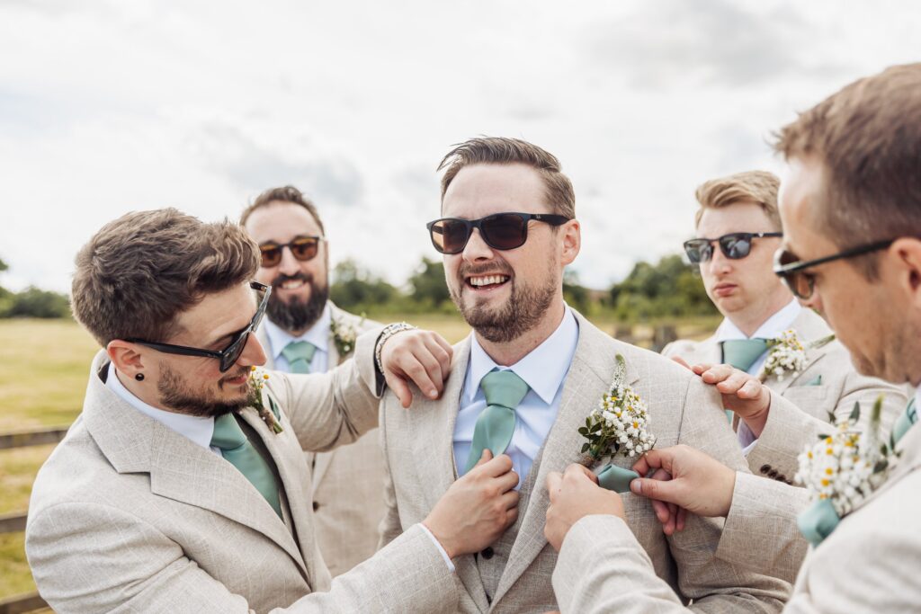 A group of men in light beige suits and teal ties adjust each other's attire outdoors at Autumn & Sam - Owen house barn