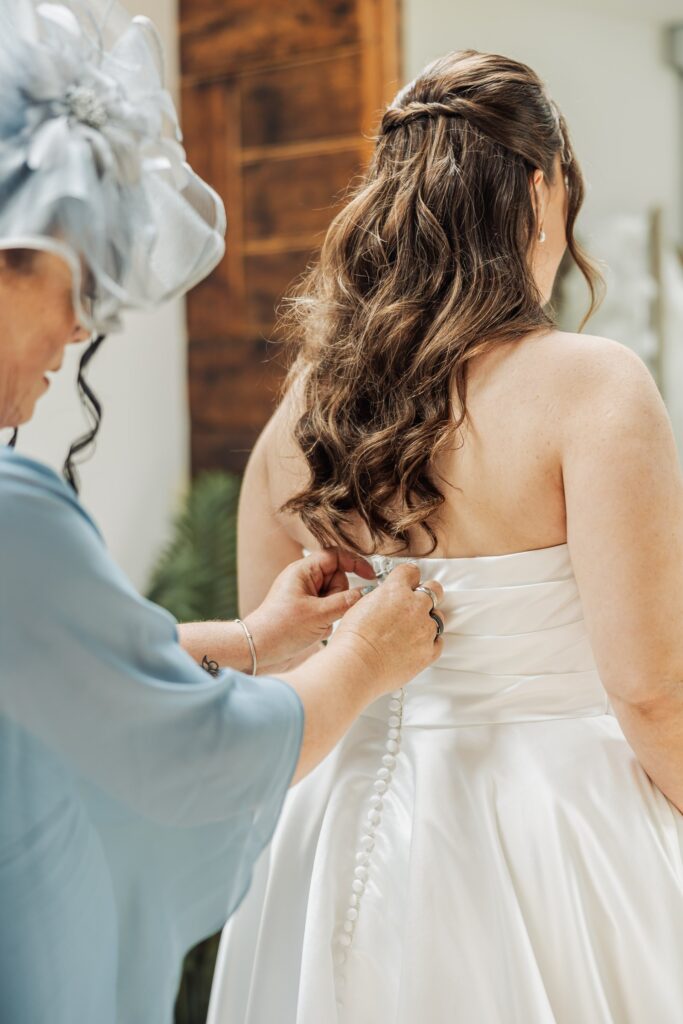An older man in a light blue top fastens the back of a bride's white gown in a warmly lit room with wooden paneling at Autumn & Sam - Owen house barn