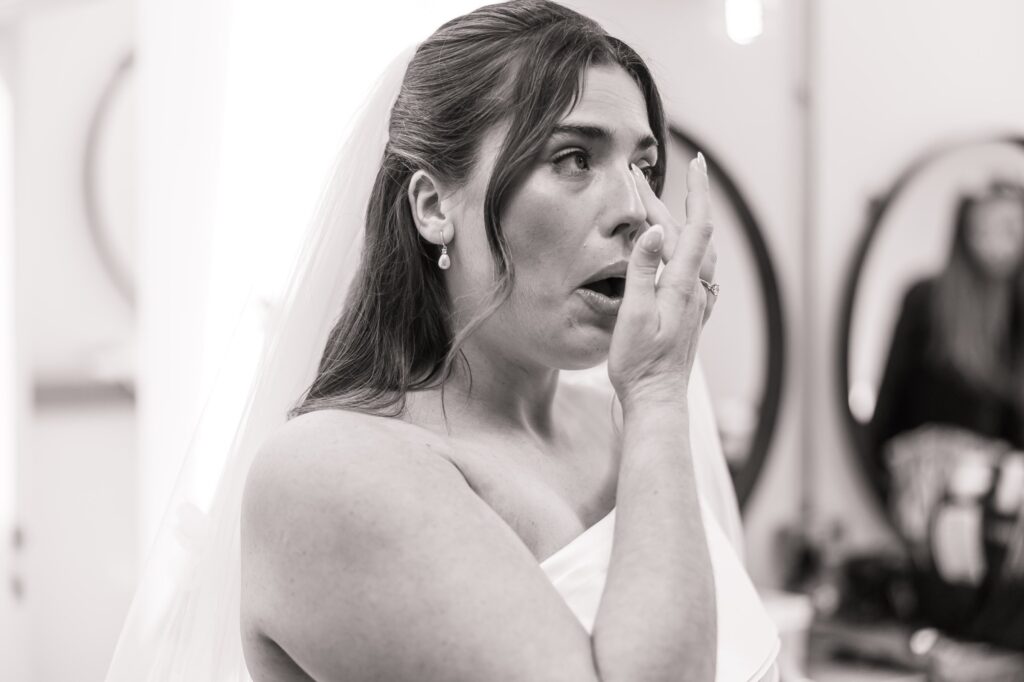 A bride in a white dress and veil adjusts her makeup, wearing pearl earrings and a ring, in a softly lit indoor setting