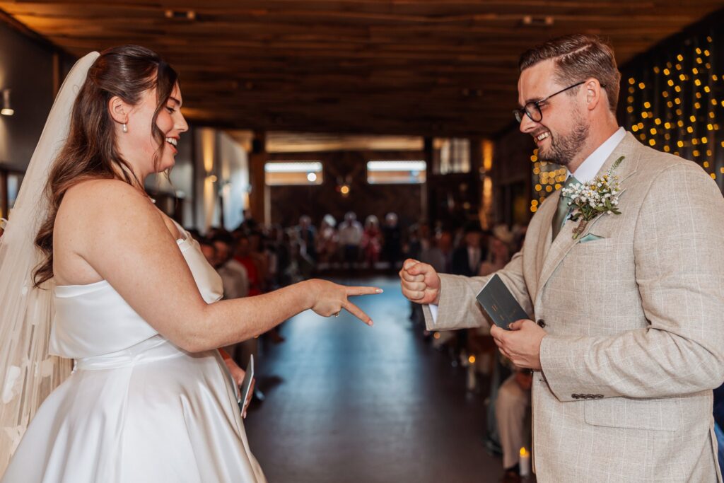 A couple in wedding attire stands facing each other in a warmly lit wooden hall, exchanging vows during a ceremony at Autumn & Sam - Owen house barn