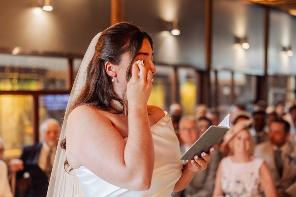 A bride in a white wedding dress and veil wipes her eye with a tissue during a wedding ceremony at Autumn & Sam - Owen house barn