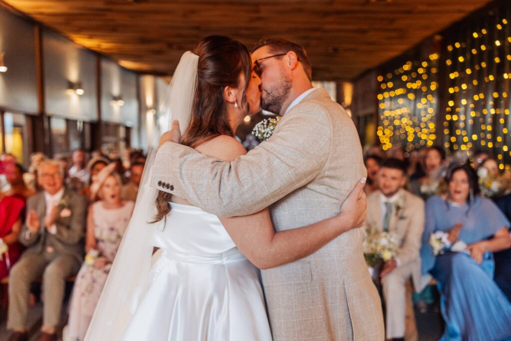 Autumn and Sam kissing at the end of the aisle in the barn at Owen House, guests and fairy lights behind them
