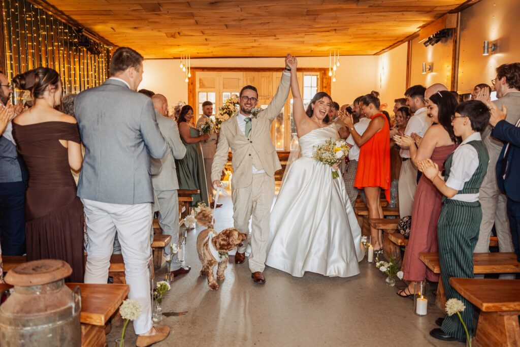 A couple in wedding attire walks down an aisle, hand in hand, with a small dog on a leash at Autumn & Sam - Owen house barn