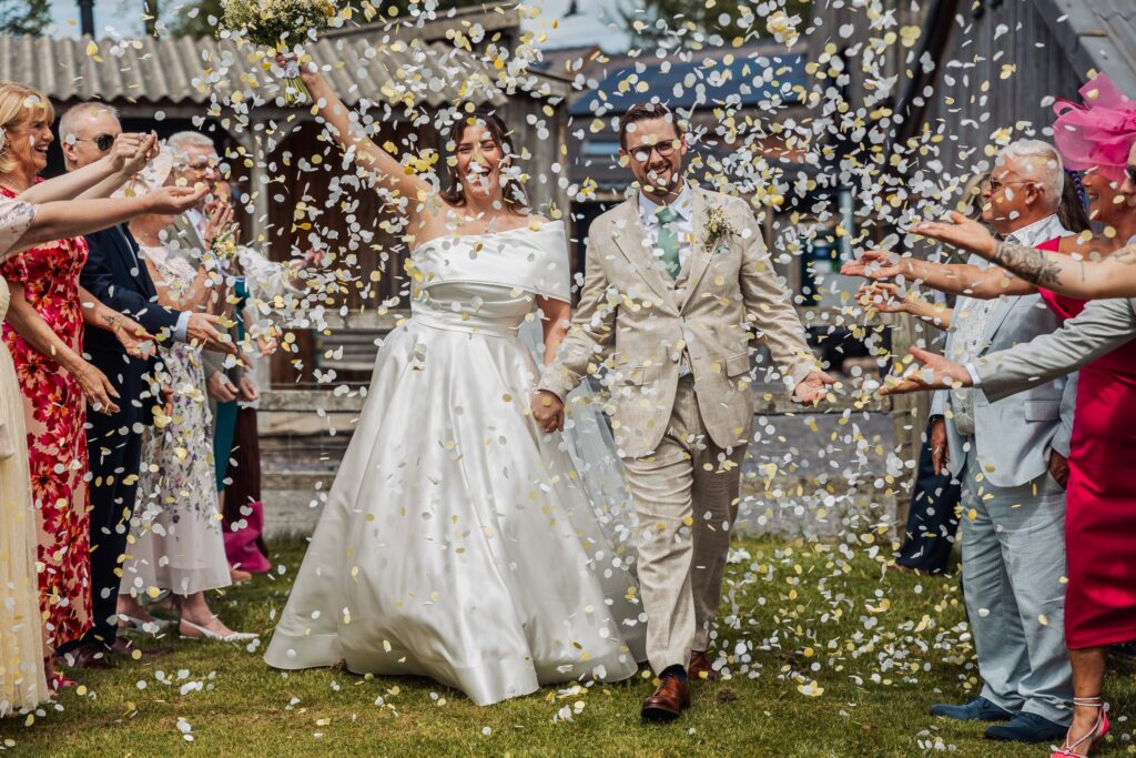 A couple in wedding attire walks through a shower of confetti, surrounded by guests throwing confetti in a rustic outdoor setting at Autumn & Sam - Owen house barn
