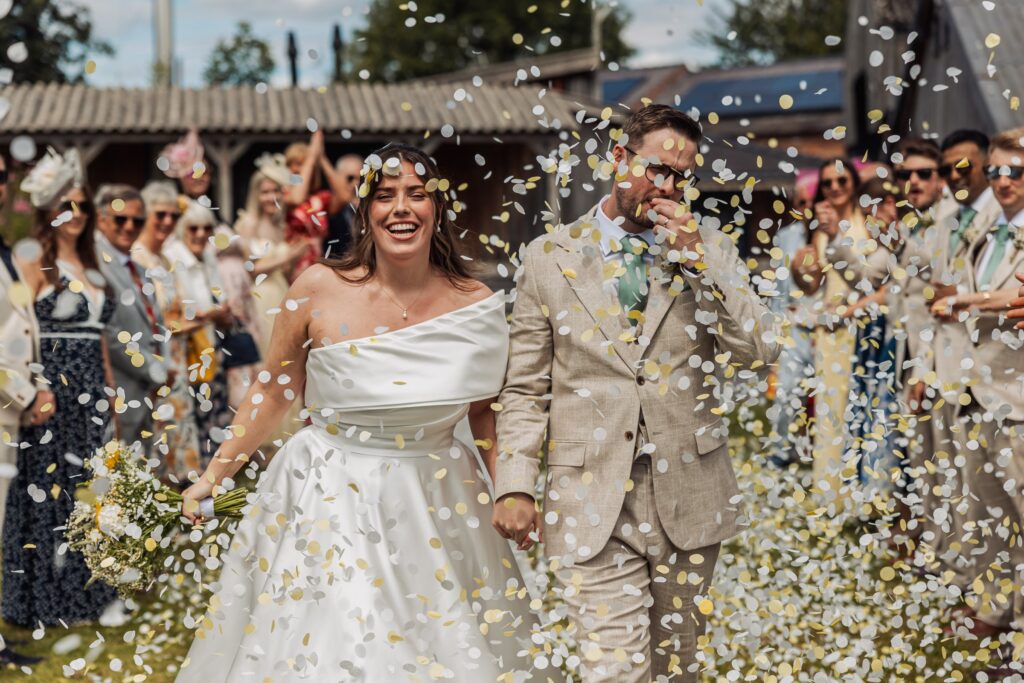 A couple in wedding attire walks through a shower of confetti, with guests in the background throwing more confetti at Autumn & Sam - Owen house barn