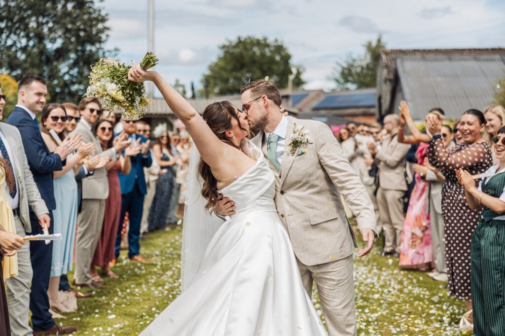 A couple kisses as the bride raises her bouquet, with guests clapping and confetti scattered on the ground at Autumn & Sam - Owen house barn