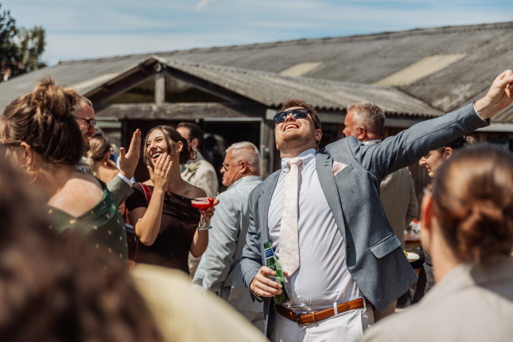 A man in a grey suit and sunglasses raises his arm in celebration, holding a bottle of beer, surrounded by laughing guests at an outdoor wedding venue