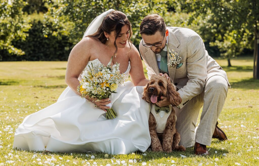 A couple in wedding attire sits on a grassy lawn, holding a bouquet and petting a small dog wearing a bow tie at Autumn & Sam - Owen house barn
