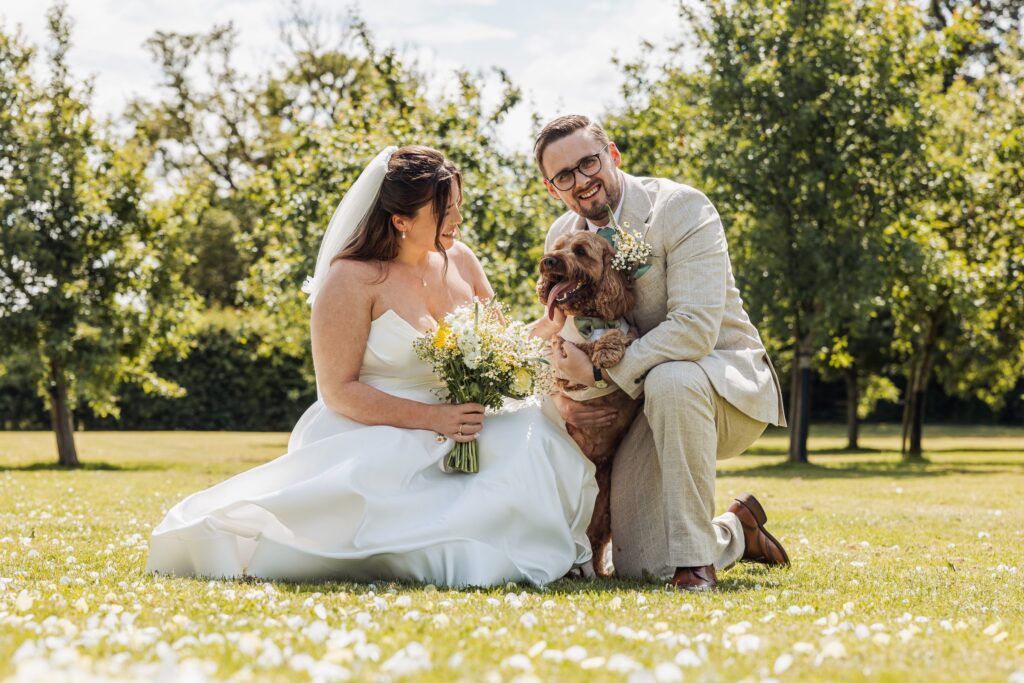 Bride and groom sitting on the daisy lawn with their cockapoo at Owen House Wedding Barn