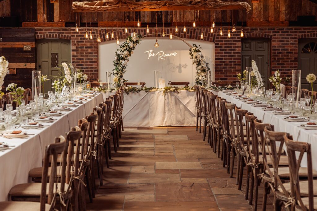 The barn at Owen House set for the wedding breakfast with long banquet tables, festoon lights and a floral arch
