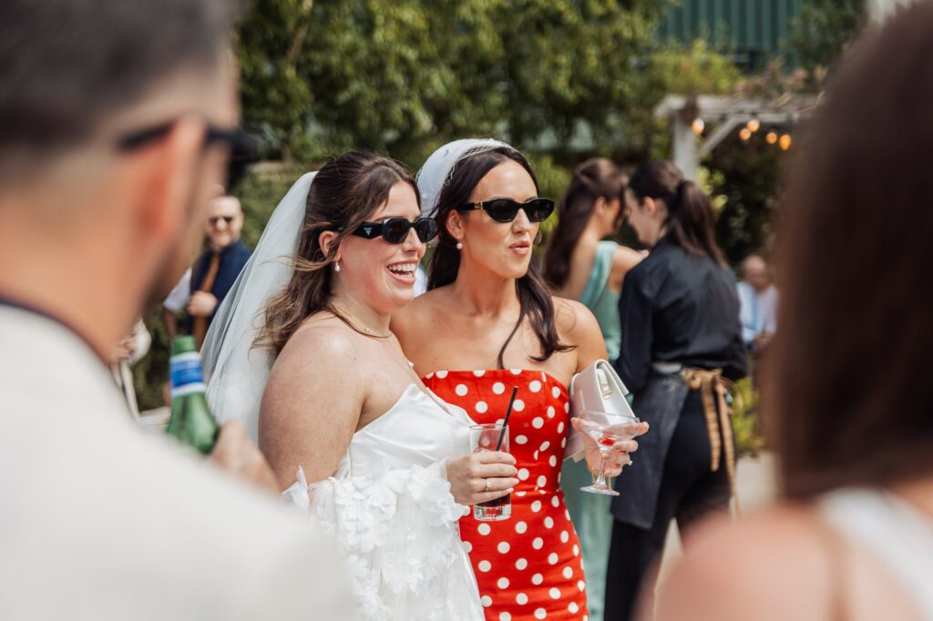 Two women in wedding attire smile and hold drinks in a garden setting with other guests in the background
