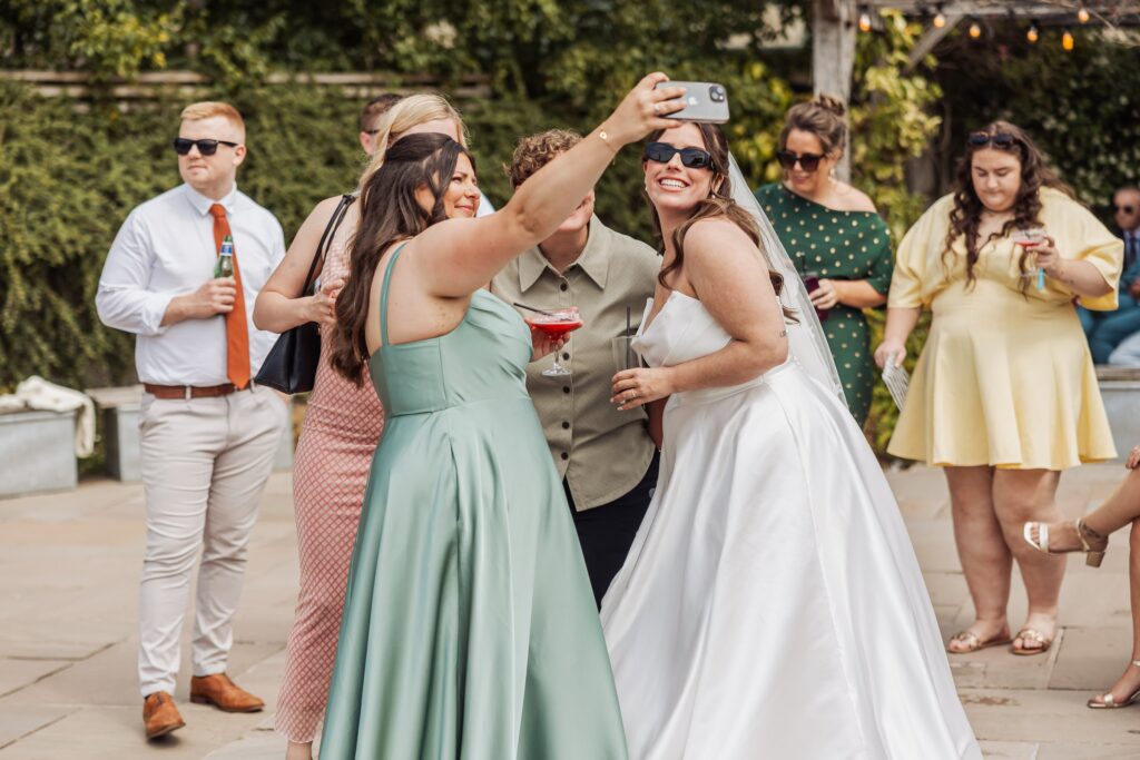 Two women in wedding attire take a selfie with a smartphone, surrounded by other guests in an outdoor setting with greenery at Autumn & Sam - Owen house barn