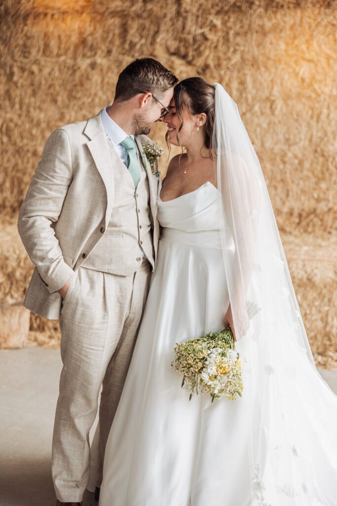 A couple in wedding attire stands close together in front of a backdrop of straw bales