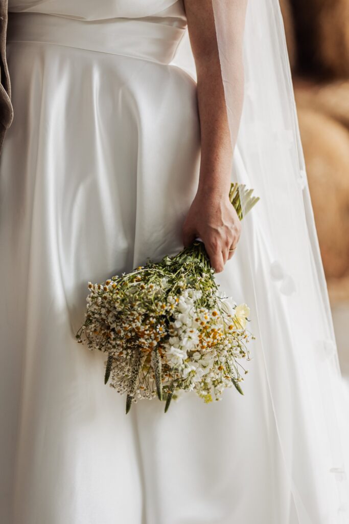 A person in a white wedding dress holds a small bouquet of white and yellow flowers at Autumn & Sam - Owen house barn