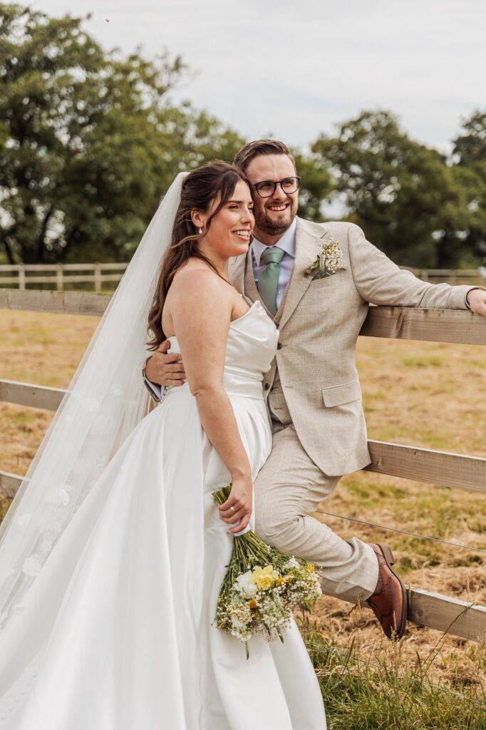 A couple in wedding attire stands close together, the woman in a white strapless gown and veil, holding a bouquet of wildflowers