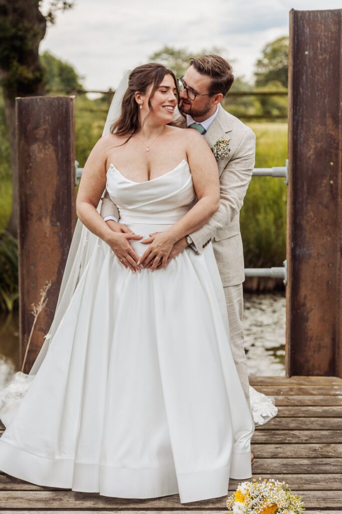A couple in wedding attire stands close together on a wooden dock, embracing at Autumn & Sam - Owen house barn