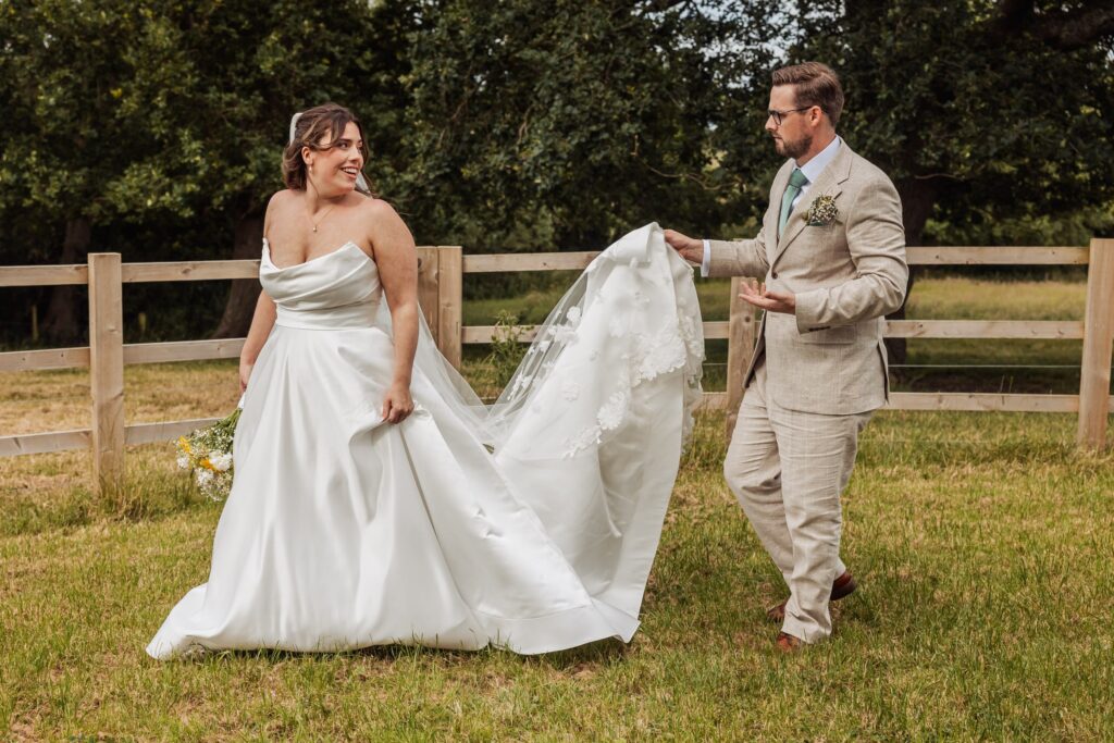 A couple in wedding attire walks in a grassy field, with the woman holding a bouquet and the man holding her dress at Autumn & Sam - Owen house barn