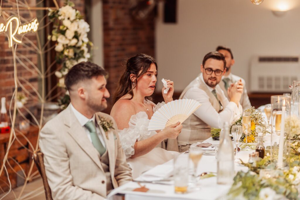 A bride in a white dress with ruffled sleeves and a groom in a light beige suit are seated at a long table at Autumn & Sam - Owen house barn