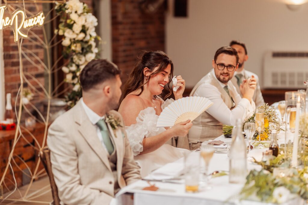 A bride in a white gown with ruffled sleeves and a man in a light beige suit with a teal tie are seated at a long table, laughing and holding a white fan