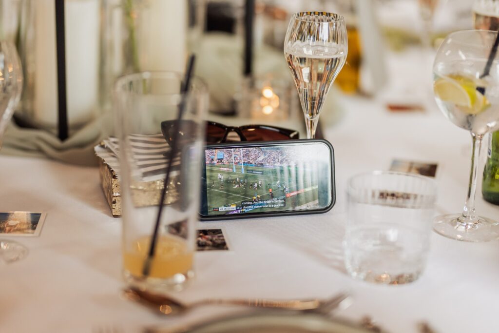 A smartphone displaying a rugby match on a table at a wedding reception, surrounded by drinks and tableware at Autumn & Sam - Owen house barn