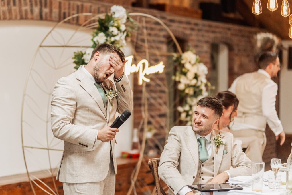 A man in a light beige suit gives a speech while another man in similar attire sits at a table with a tablet at Autumn & Sam - Owen house barn