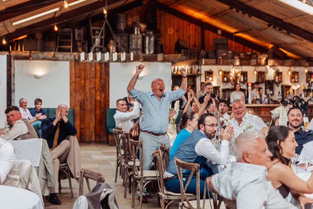 A man raises his arms in celebration during a wedding reception, surrounded by guests clapping and smiling in a rustic wooden room at Autumn & Sam - Owen house barn