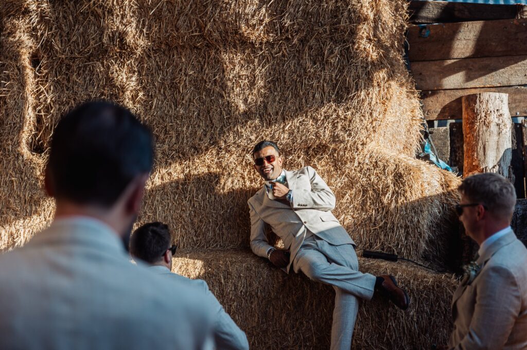 A man in a light grey suit and sunglasses lies on a large stack of hay, smiling and adjusting his tie at Autumn & Sam - Owen house barn