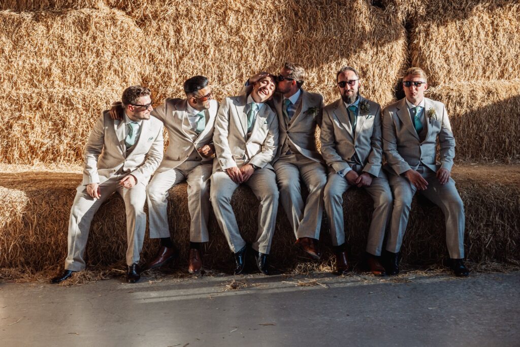 A group of men in light grey suits and teal ties sit on hay bales, smiling and appearing relaxed at Autumn & Sam - Owen house barn