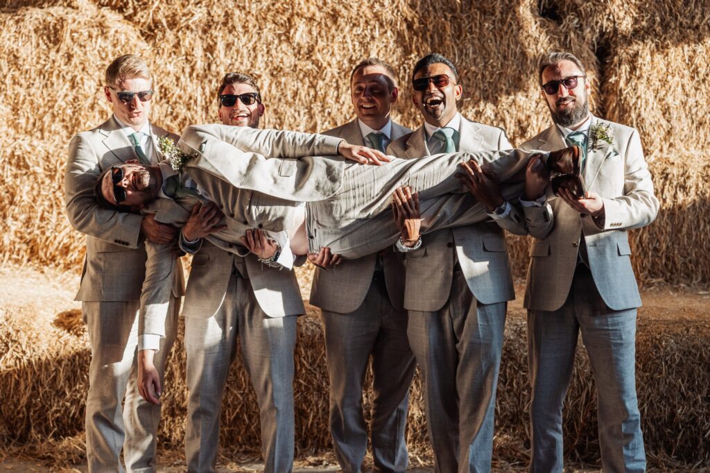 A group of men in matching light grey suits and sunglasses hold another man upside down, smiling and enjoying themselves against a backdrop of straw bales