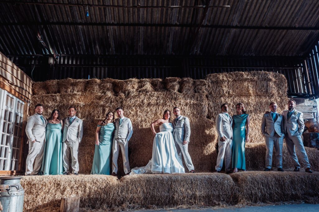A wedding party in formal attire stands in a rustic barn setting with hay bales in the background, bathed in natural light at Autumn & Sam - Owen house barn
