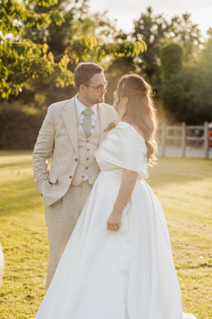 A couple in wedding attire stands close together in a grassy outdoor setting with trees and a white fence in the background at Autumn & Sam - Owen house barn