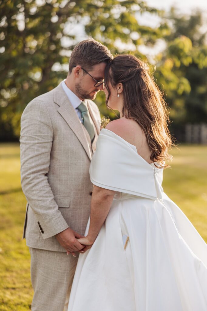 A couple in wedding attire stands close together, holding hands, outdoors with greenery and sunlight in the background