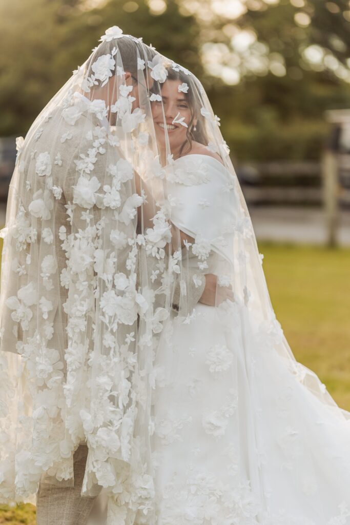 A couple in wedding attire embrace under a floral veil, with sunlight and greenery in the background at Autumn & Sam - Owen house barn