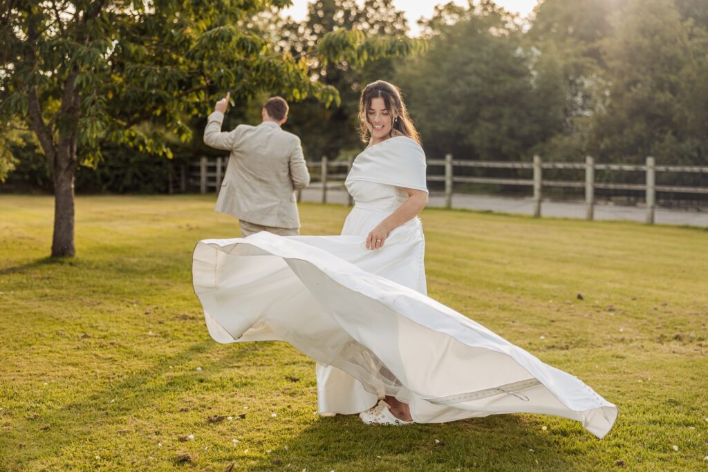 A bride in a flowing white wedding dress twirls in a grassy field, with a man in a light-coloured suit pointing in the background