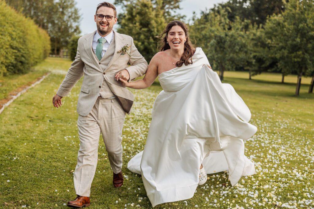 Bride and groom running hand in hand across the daisy-covered lawn at Owen House Wedding Barn