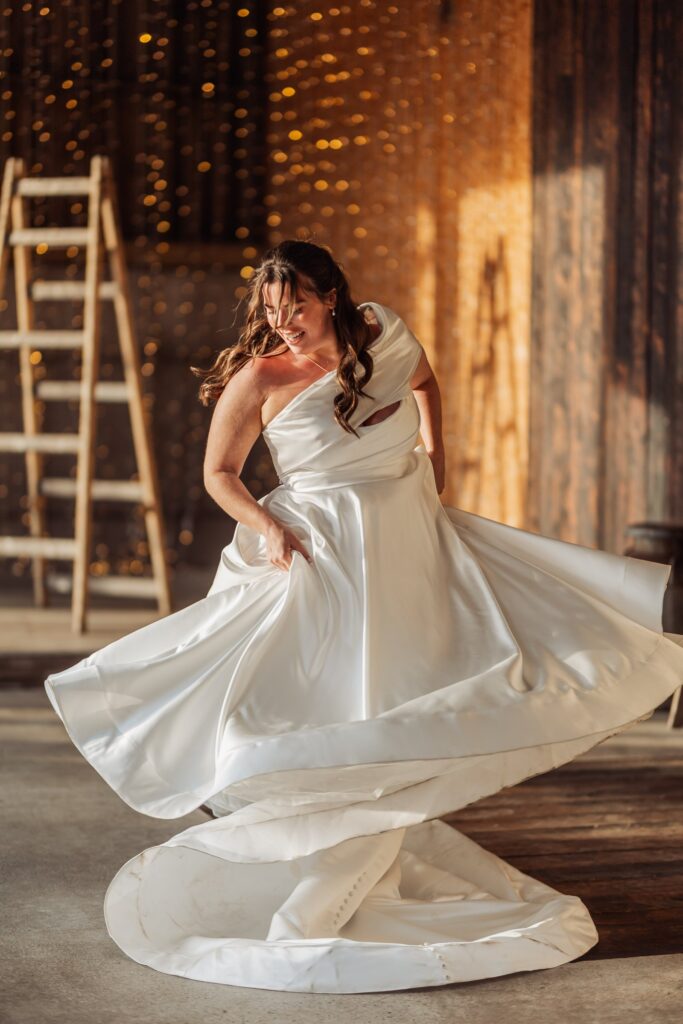 A woman in a flowing white wedding dress twirls, creating a dramatic swirl of fabric at Autumn & Sam - Owen house barn