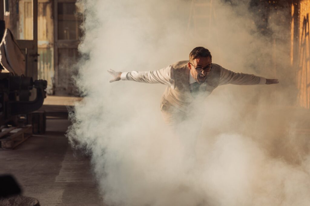 A man in a beige suit and glasses is surrounded by a cloud of white powder or smoke in an industrial or workshop setting at Autumn & Sam - Owen house barn