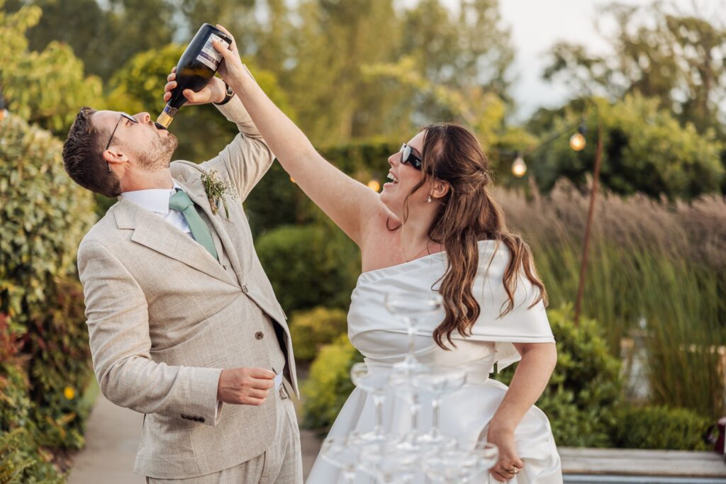 A couple in wedding attire celebrates outdoors, with the woman holding a bottle of champagne above her head and the man drinking at Autumn & Sam - Owen house barn