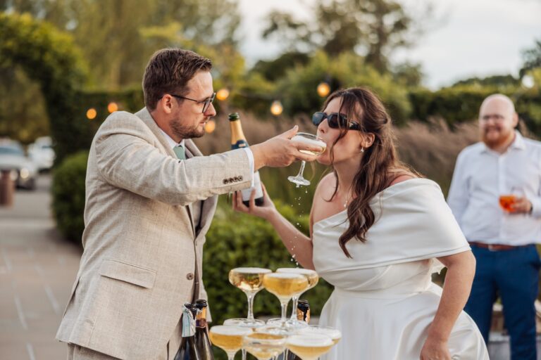 Groom pouring champagne over a tower of glasses while the bride drinks from one, sunglasses on