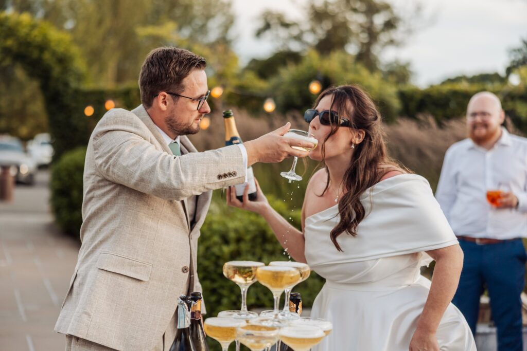 Groom pouring champagne down a champagne tower while the bride drinks from the top glass