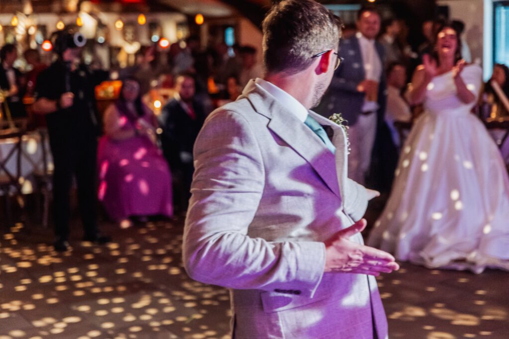 A man in a light grey suit dances on a patterned dance floor, while a woman in a white polka dot dress claps in the background at Autumn & Sam - Owen house barn