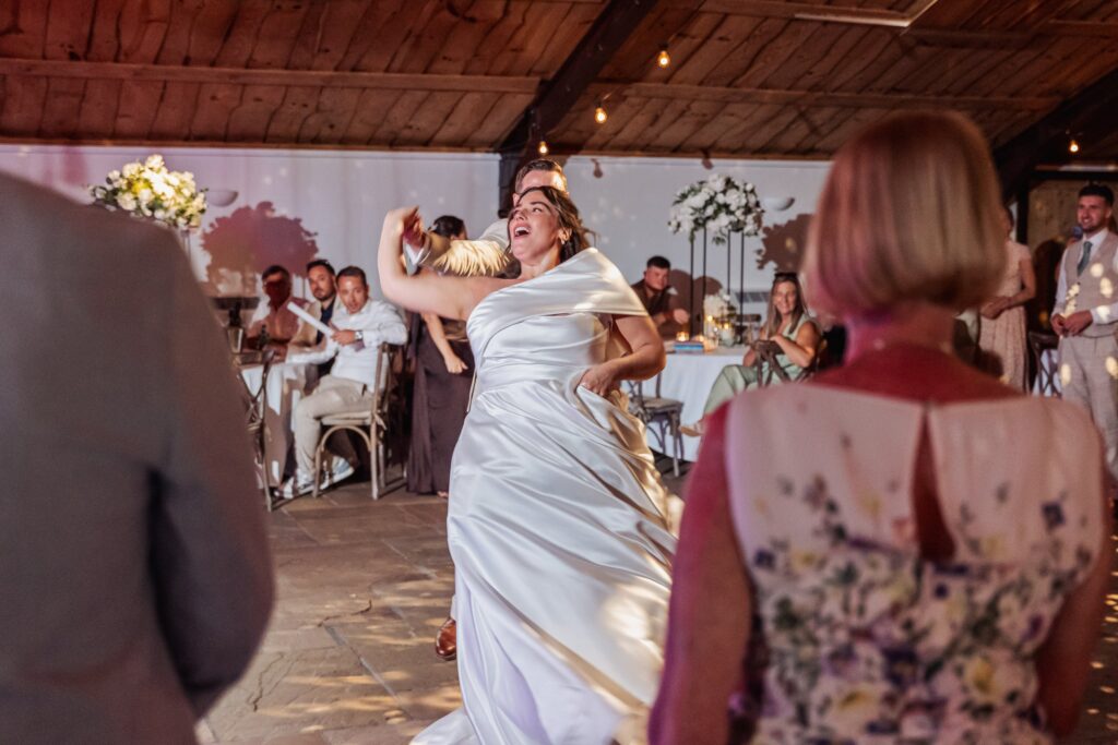 A couple dancing mid-movement on a floor, with guests seated at tables in a warm wooden room with fairy lights at Autumn & Sam - Owen house barn