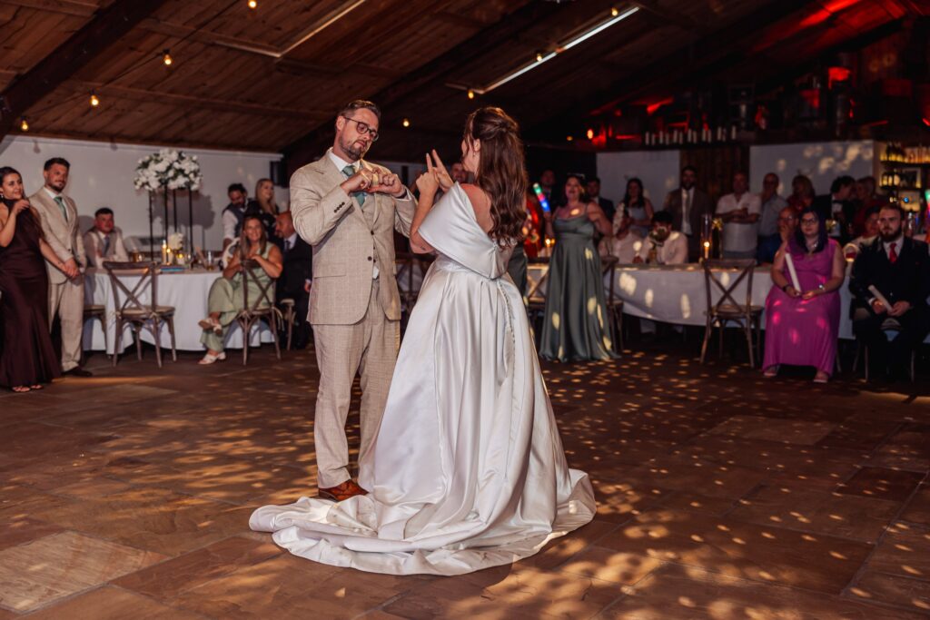 A couple dancing on a wooden floor in a warmly lit room with fairy lights, surrounded by seated guests at Autumn & Sam - Owen house barn