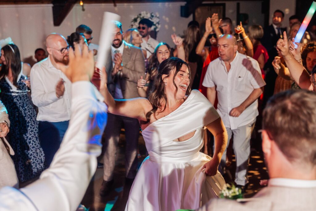 A bride in a white dress dances energetically on a dance floor surrounded by guests raising their hands in celebration at Autumn & Sam - Owen house barn